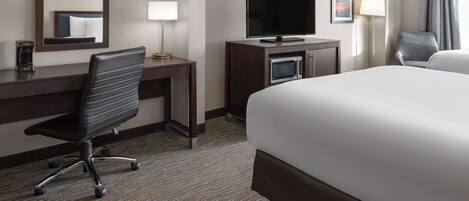 A hotel room with a large bed, a desk with a flat-screen TV, a chair, a coffee maker, and a lamp.