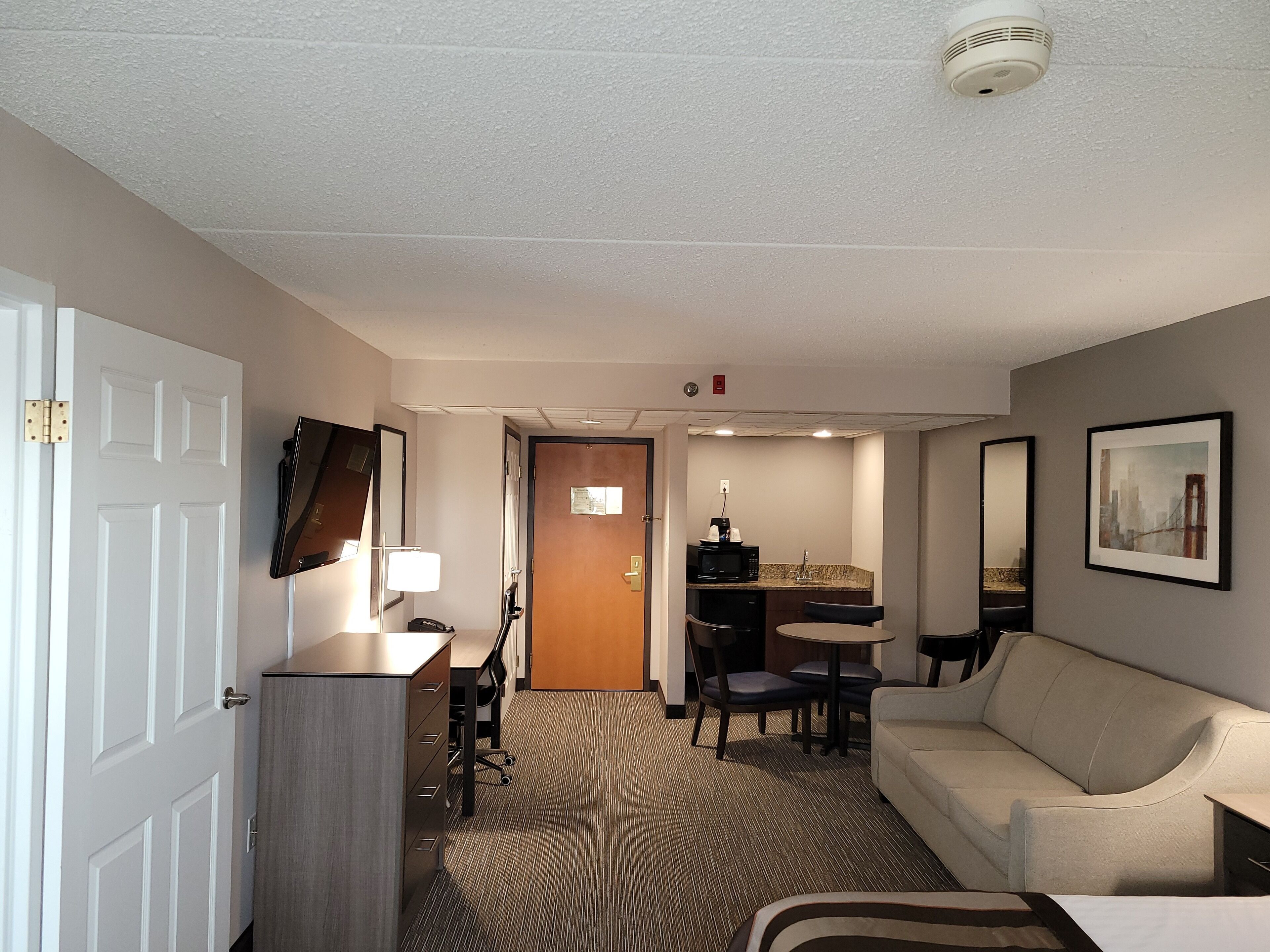 A hotel room with a desk, chair, sofa, and a small kitchenette.