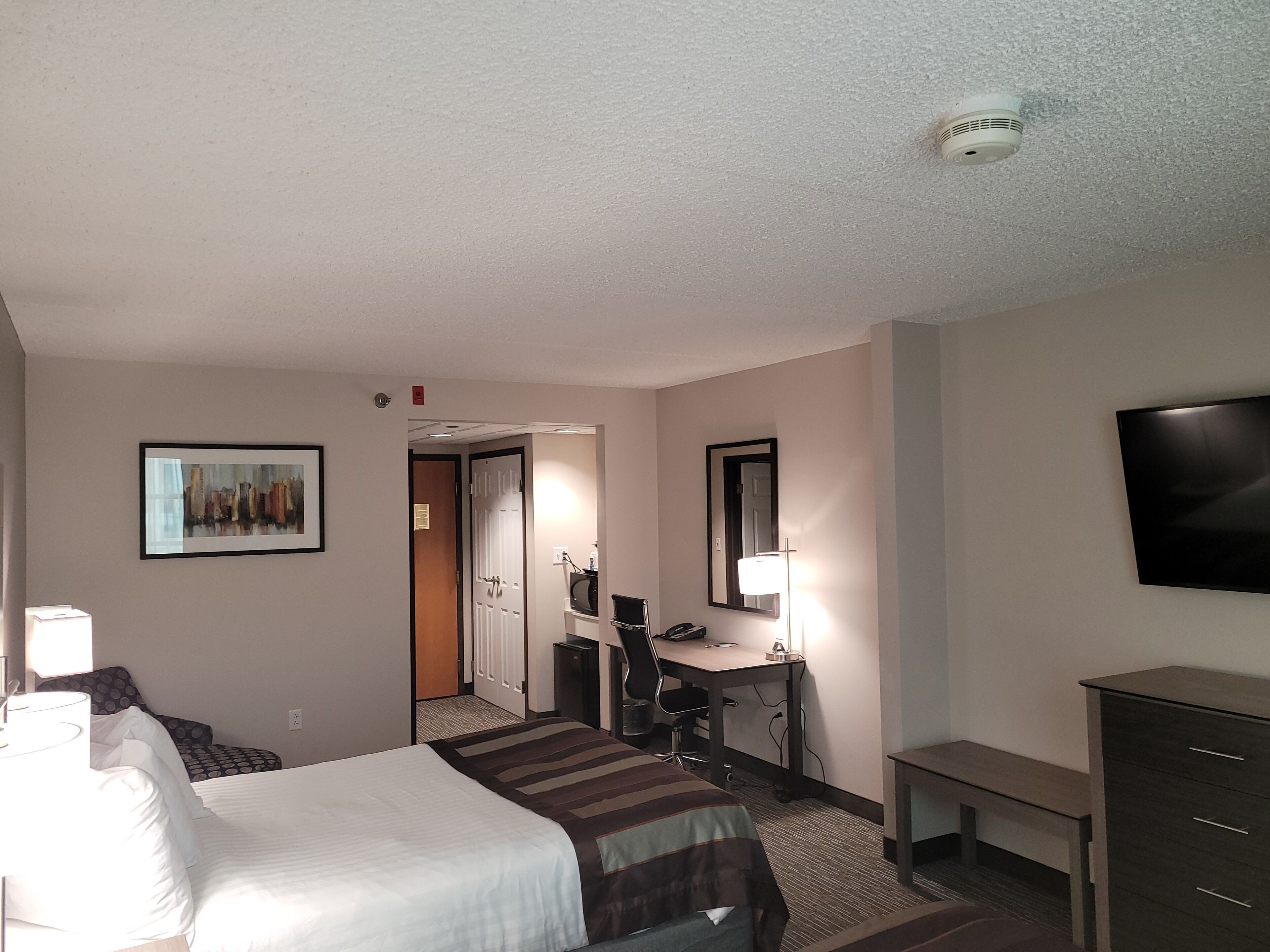 A hotel room with a bed, desk, chair, lamp, TV, and a painting on the wall.