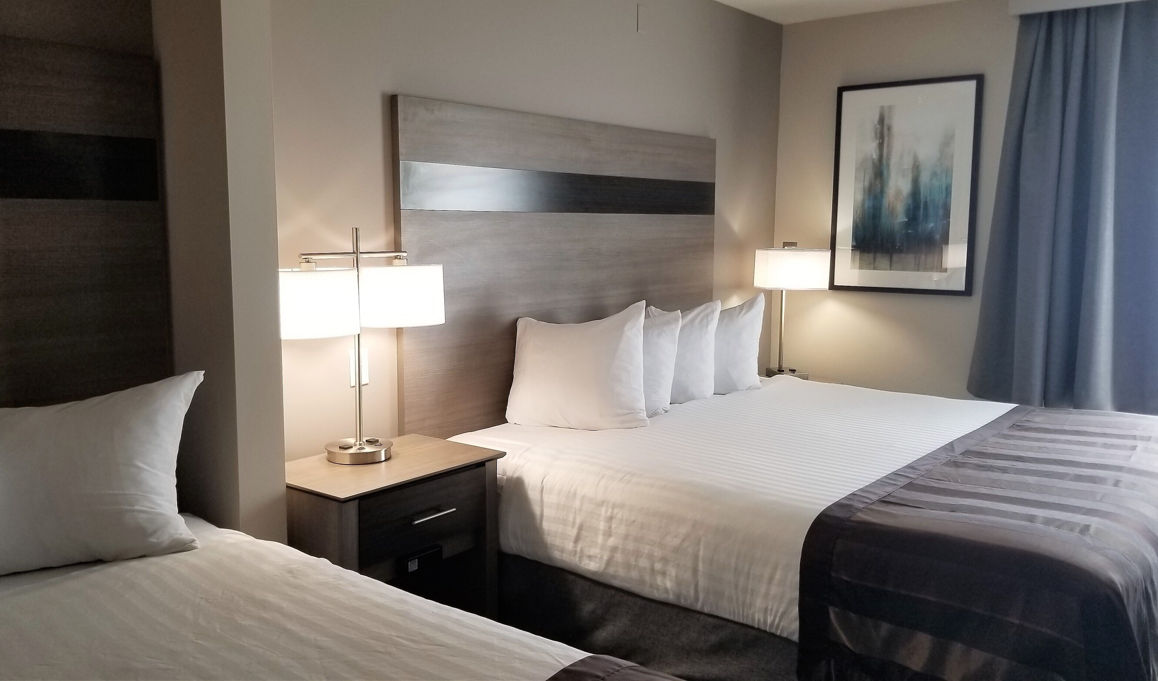 A hotel room with two beds, a nightstand with a lamp, a painting on the wall, and a window with curtains.