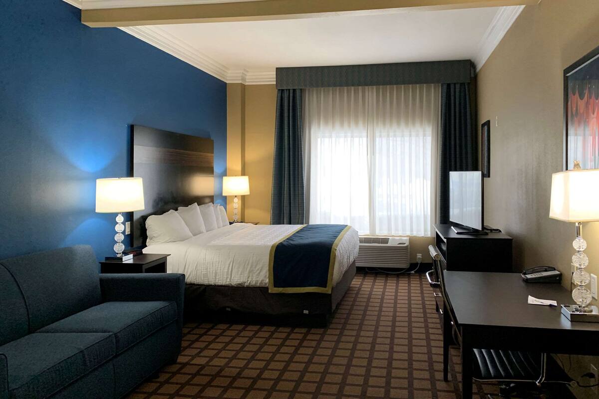 A hotel room with a bed, two bedside tables with lamps, a desk with a chair, a couch, and a large window with curtains.