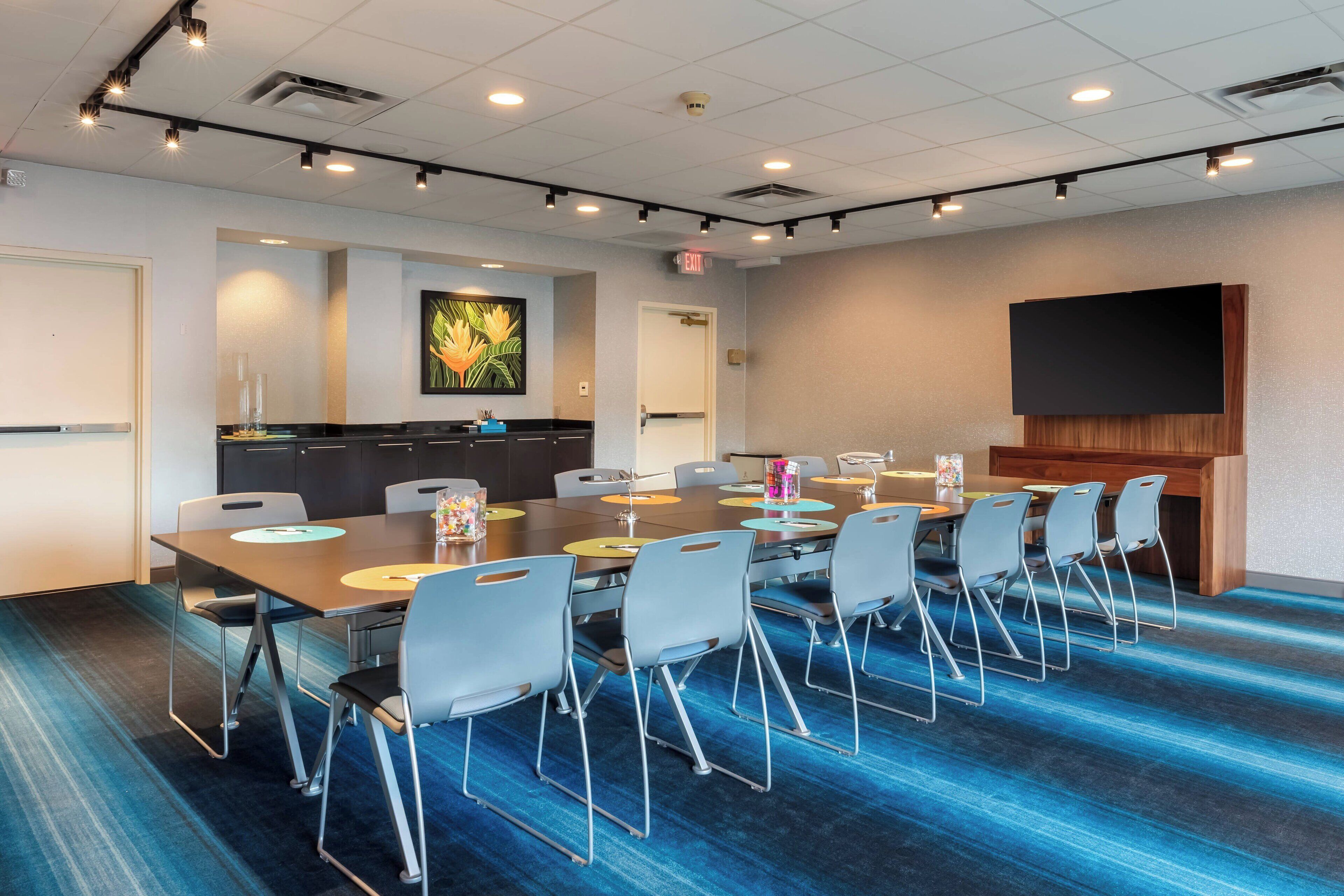 A conference room with a long table, chairs, a projector screen, and a kitchen area.