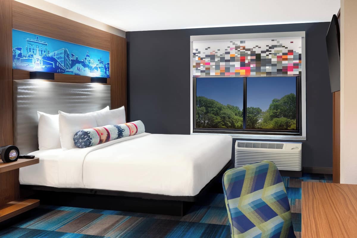 Ein Hotelzimmer mit Bett, Nachttisch, Sessel, großem Fenster mit Ausblick und einem Wandbild.