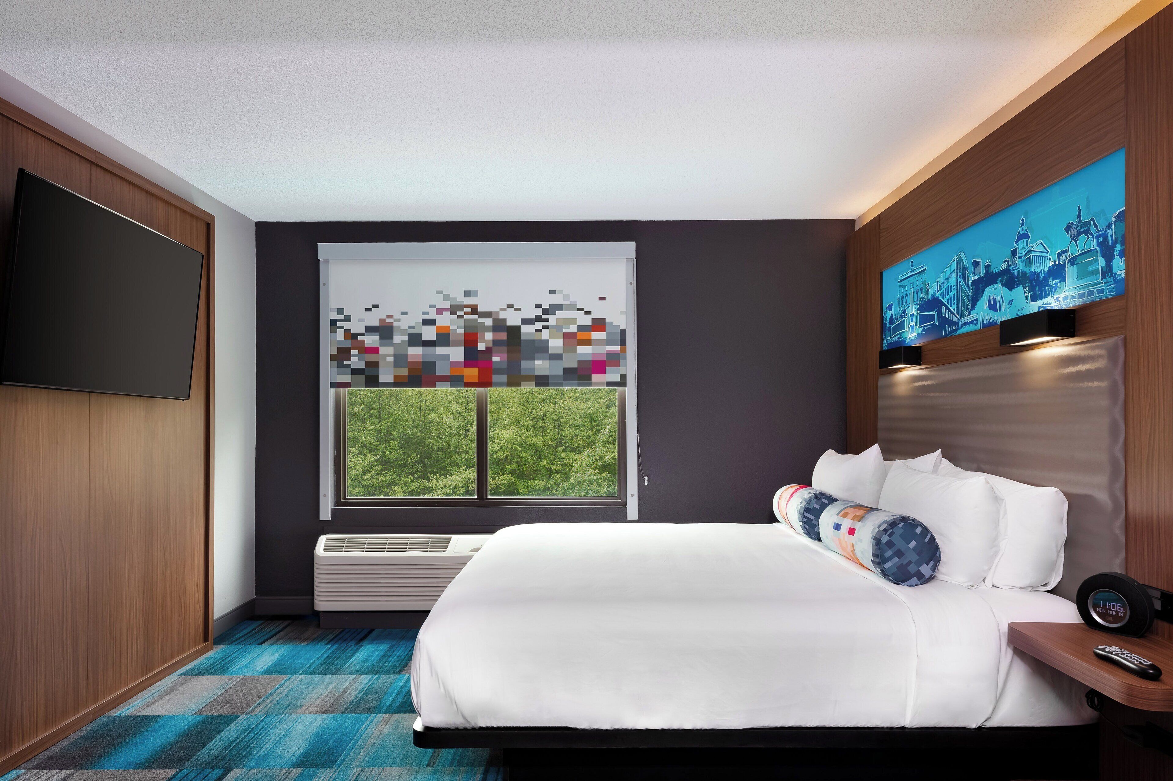 A modern hotel room with a large bed, a flat-screen TV, a window with a view of greenery, and a digital art piece on the wall.