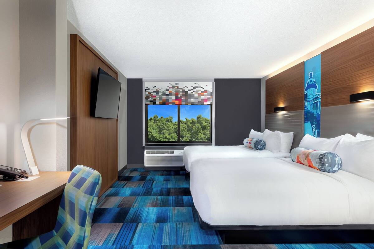 Ein modernes Hotelzimmer mit einem großen Bett, einem Schreibtisch mit Stuhl, einem Flachbildfernseher und einem Fenster mit Blick auf Bäume.