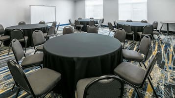 A conference room with a large round table surrounded by chairs, a projector screen, and a large window with blinds.