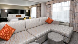 Et moderne hotelværelse med en stor hjørnesofa, en rund skammel, en seng med mange puder og et stort vindue med gardiner.