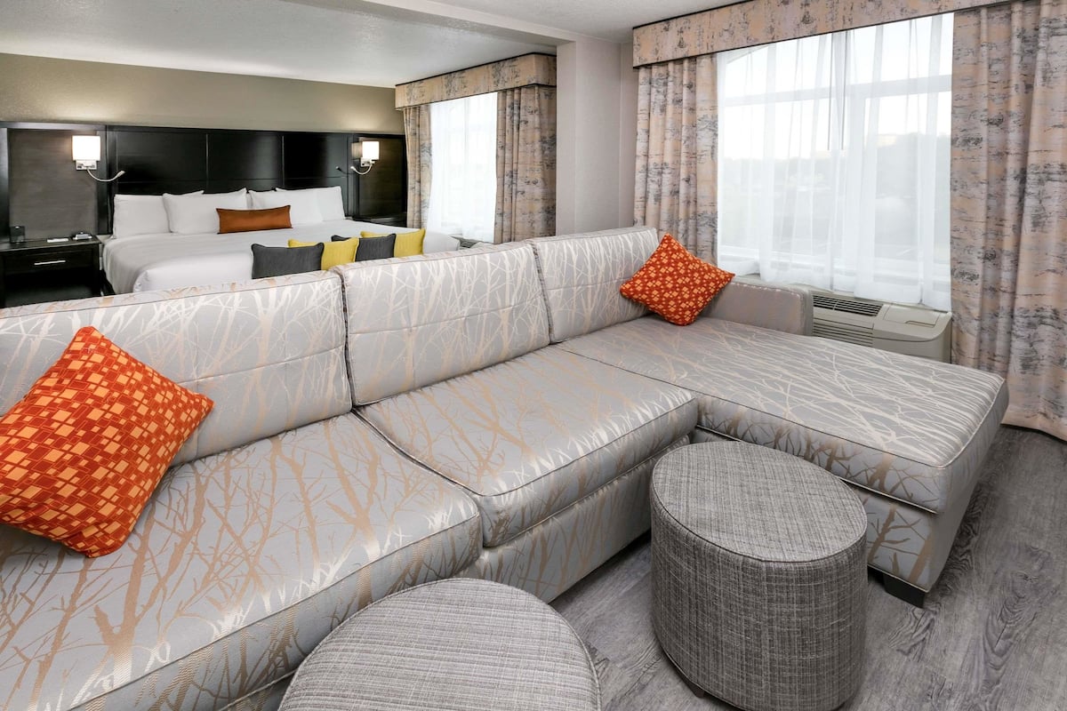 A modern hotel room with a large sectional sofa, a round ottoman, a bed with multiple pillows, and a large window with curtains.