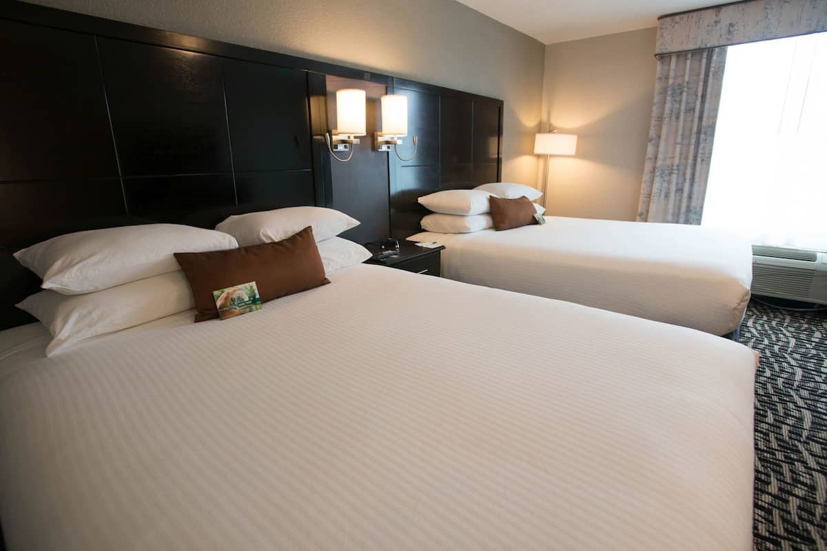 A hotel room with two beds, a dark headboard, wall-mounted lamps, a nightstand, and a window with curtains.