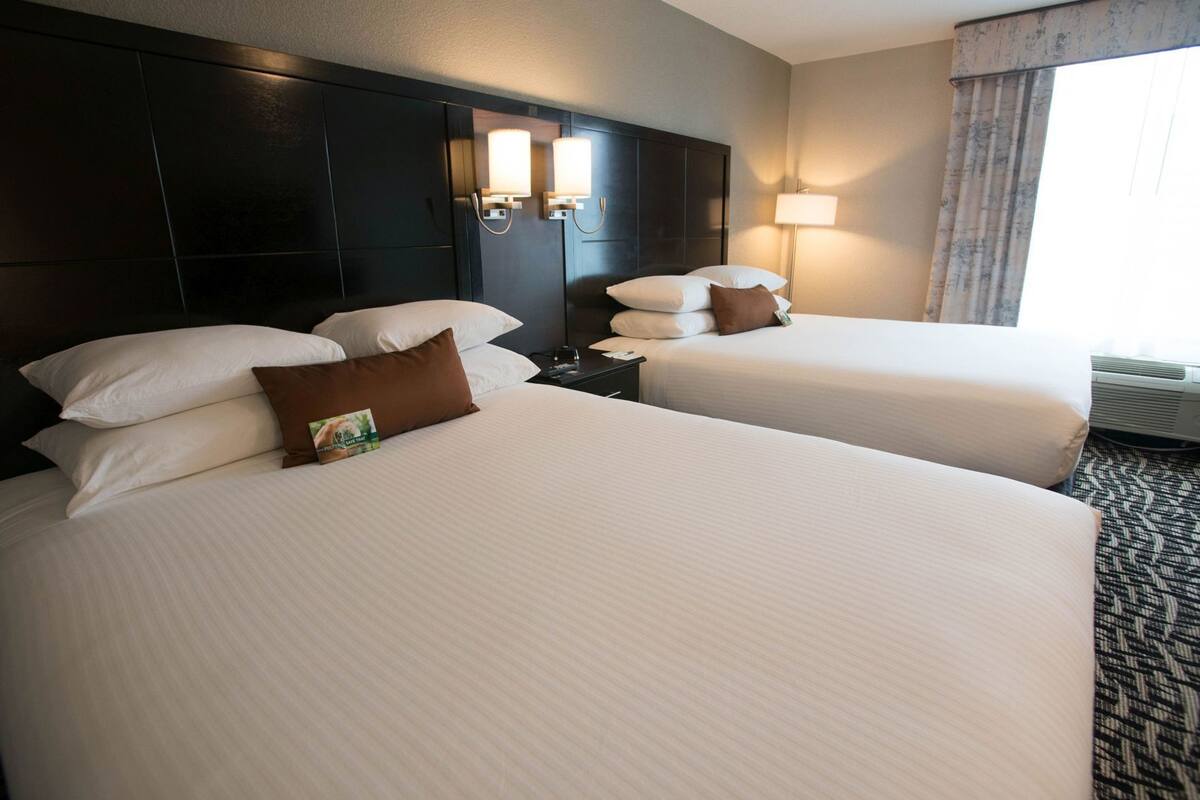A hotel room with two beds, a dark headboard, wall-mounted lamps, a nightstand, and a window with curtains.