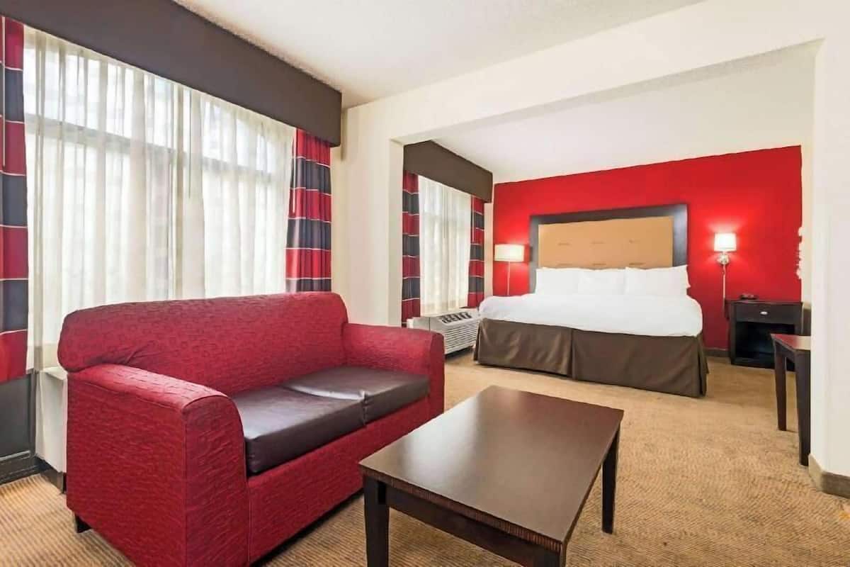 A hotel room with a red sofa, a bed with a white headboard, a small table, and a nightstand with a lamp.