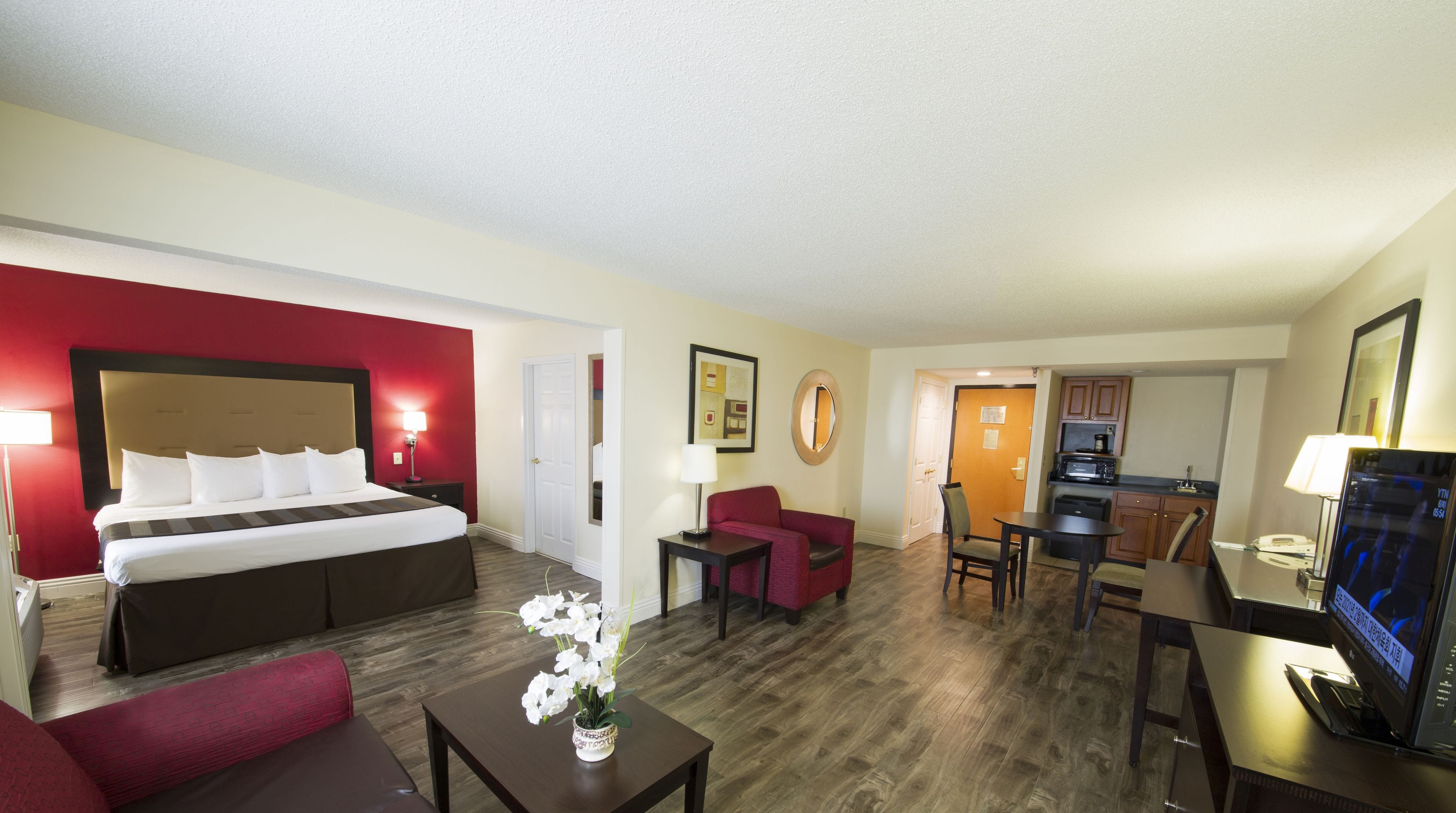 A hotel room with a bed, a desk, a chair, a small table with a vase of flowers, a television, and a kitchen area.