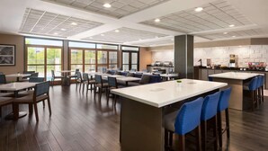Uma zona de refeições moderna com várias mesas e cadeiras, uma ilha central com banquetas e uma área de cozinha ao fundo.