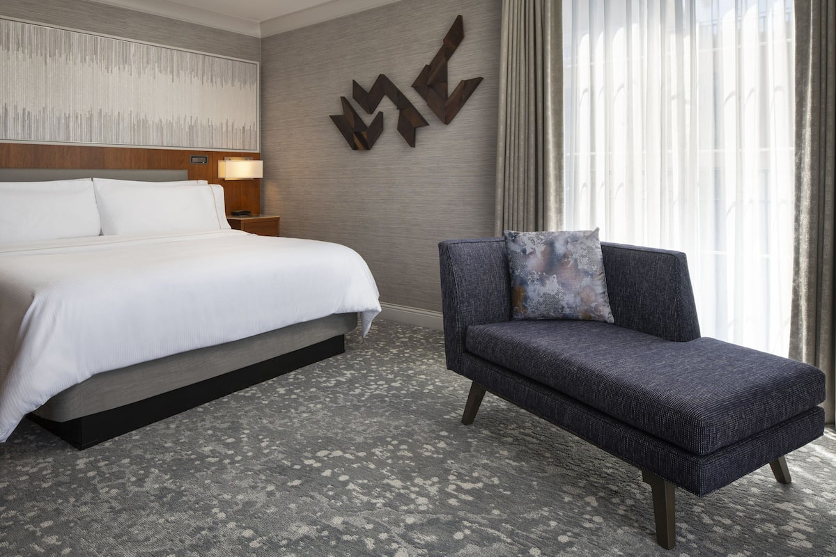 A hotel room with a large bed, a sofa, a wall-mounted artwork, and a window with curtains.
