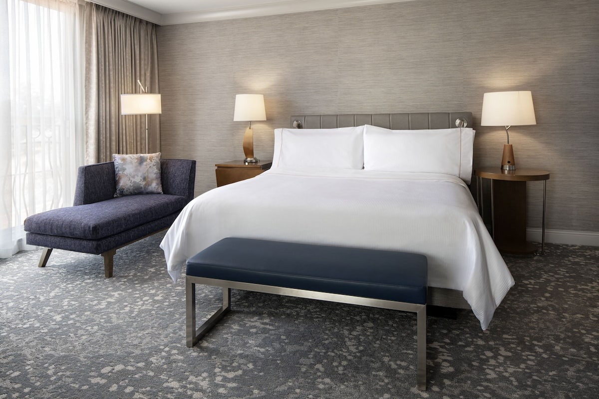 A hotel room with a large bed, two bedside tables with lamps, a chair, and a bench.
