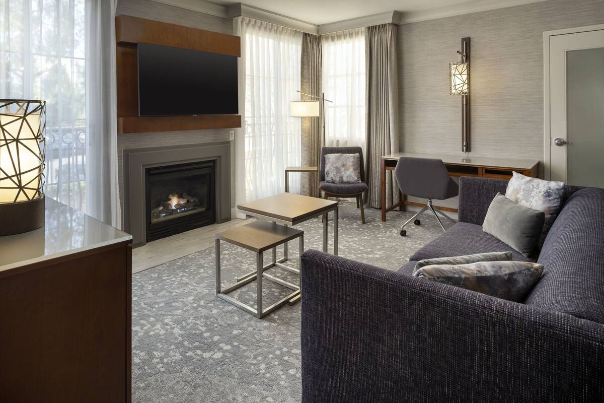 A modern hotel room with a fireplace, a flat-screen TV, a sofa, a chair, and a desk.