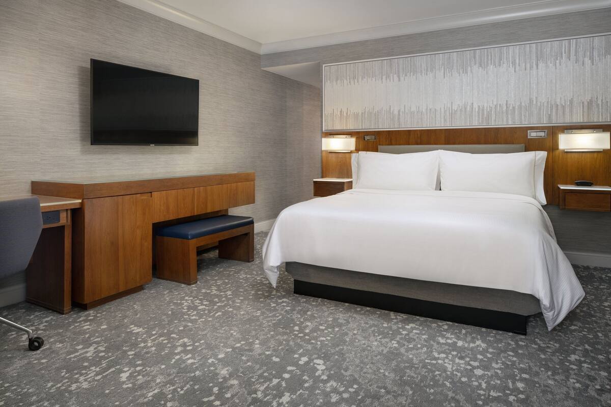 A hotel room with a large bed, a desk, a chair, a flat-screen TV mounted on the wall, and two bedside lamps.