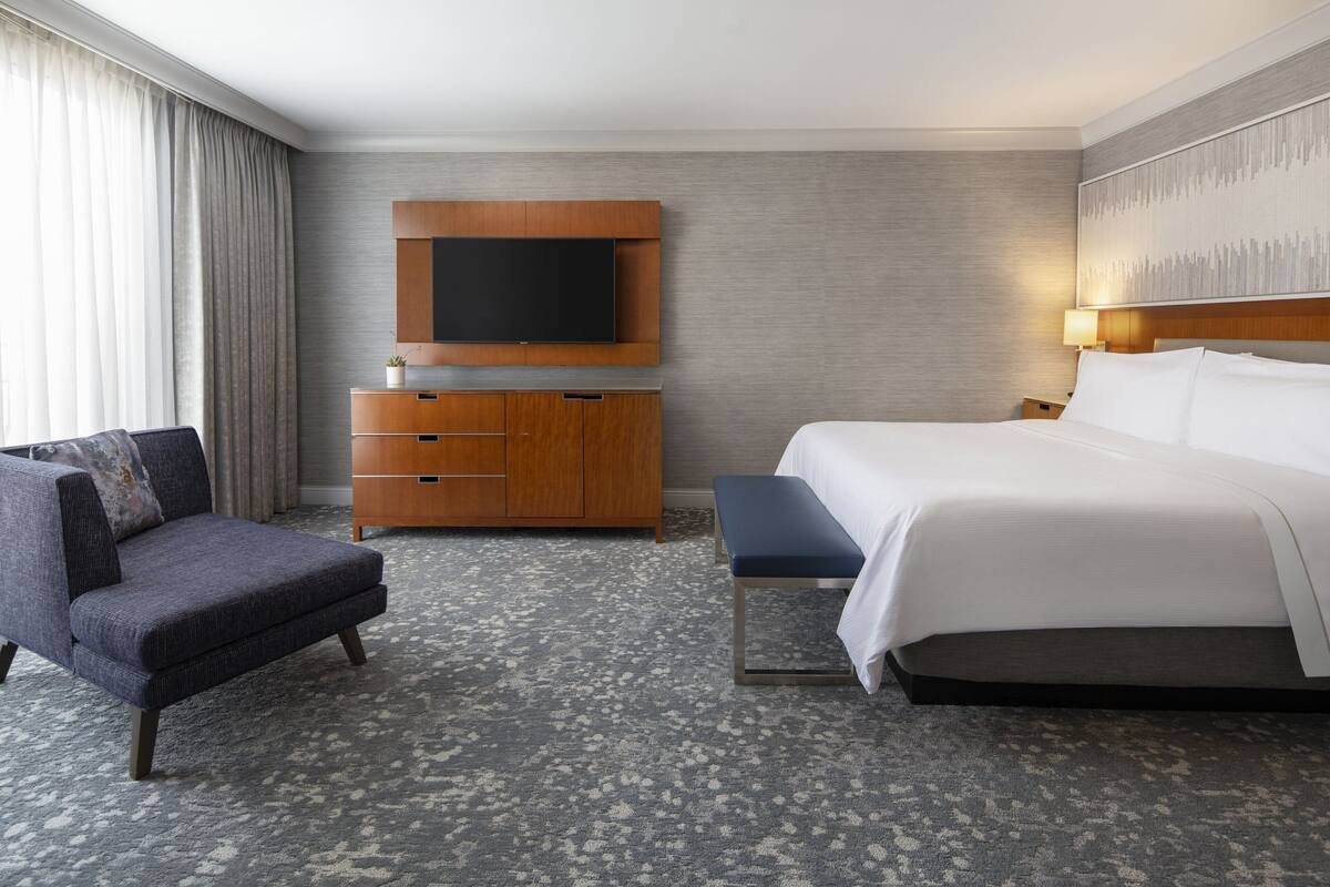 A hotel room with a bed, a TV mounted on the wall, a wooden cabinet, a chair, and a bench.