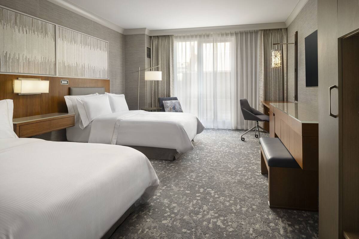A hotel room with two beds, a desk, a chair, and a large window with curtains.