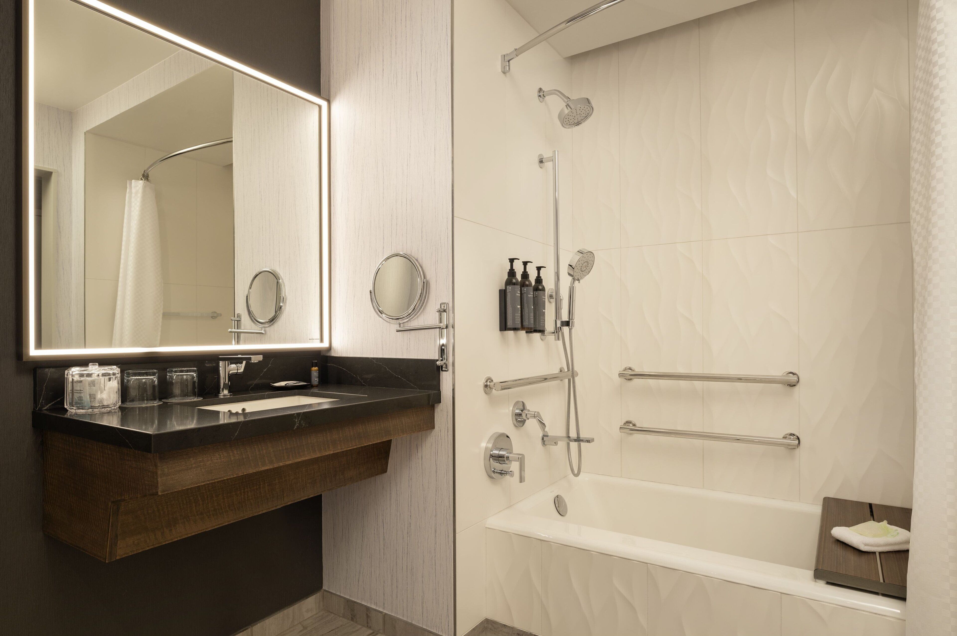 A modern bathroom with a large mirror, a vanity with a dark countertop, a bathtub, and a shower with a rain showerhead.