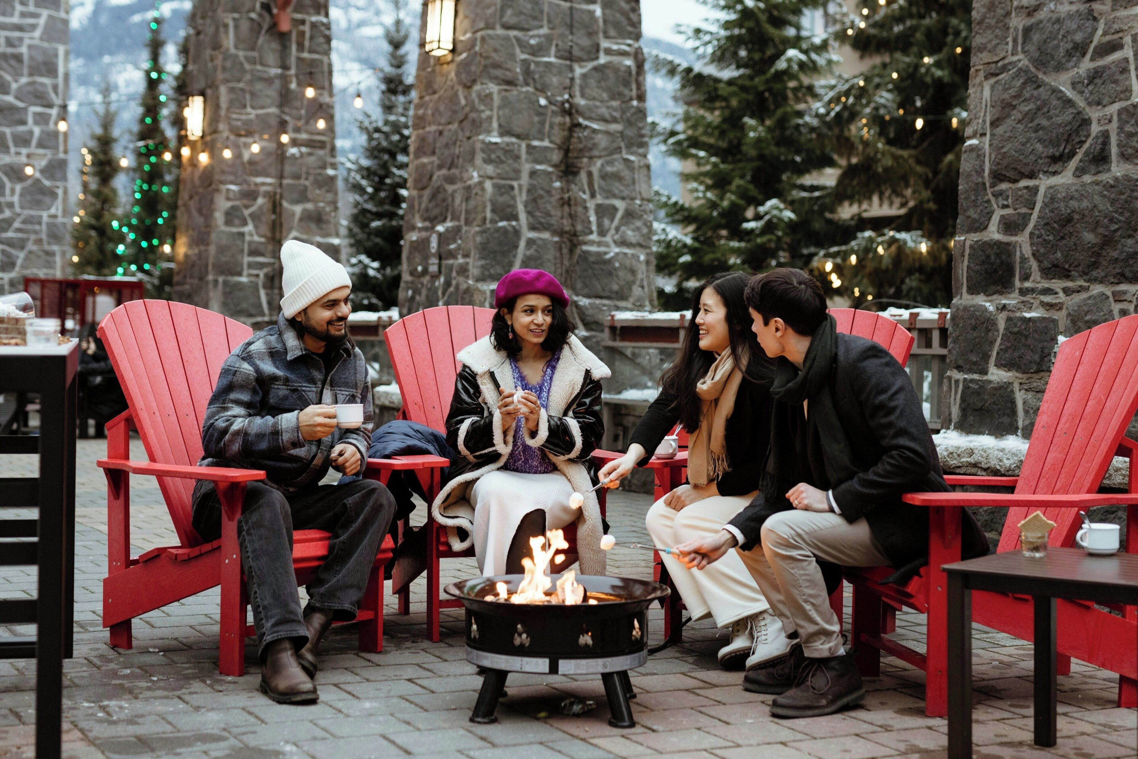 A group of people sitting on red chairs around a fire pit in an outdoor setting with stone walls and Christmas decorations.