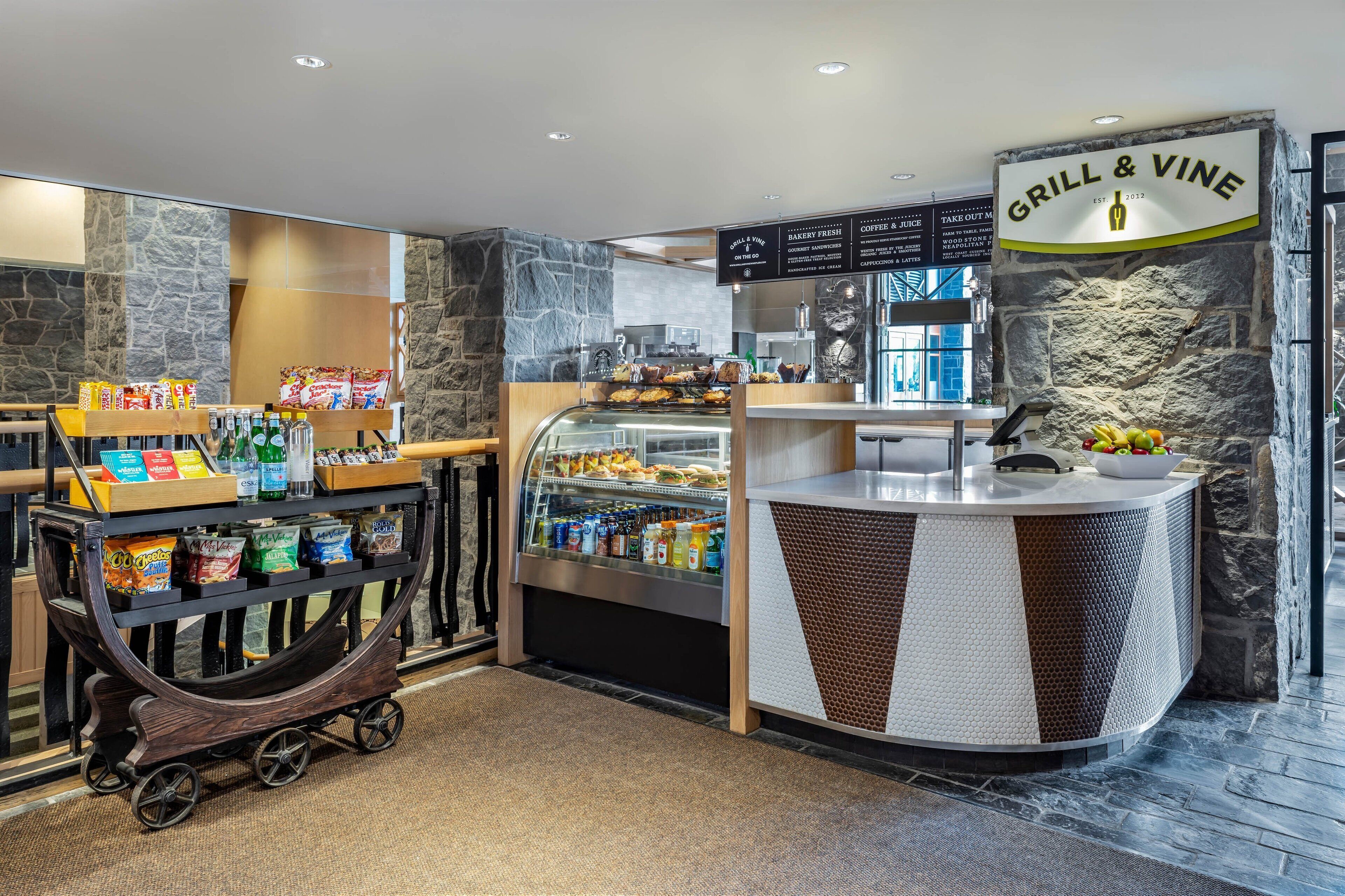 Mostrador de restaurante con pared de piedra de fondo, un tablero de menú y diversos alimentos y bebidas en exhibición.