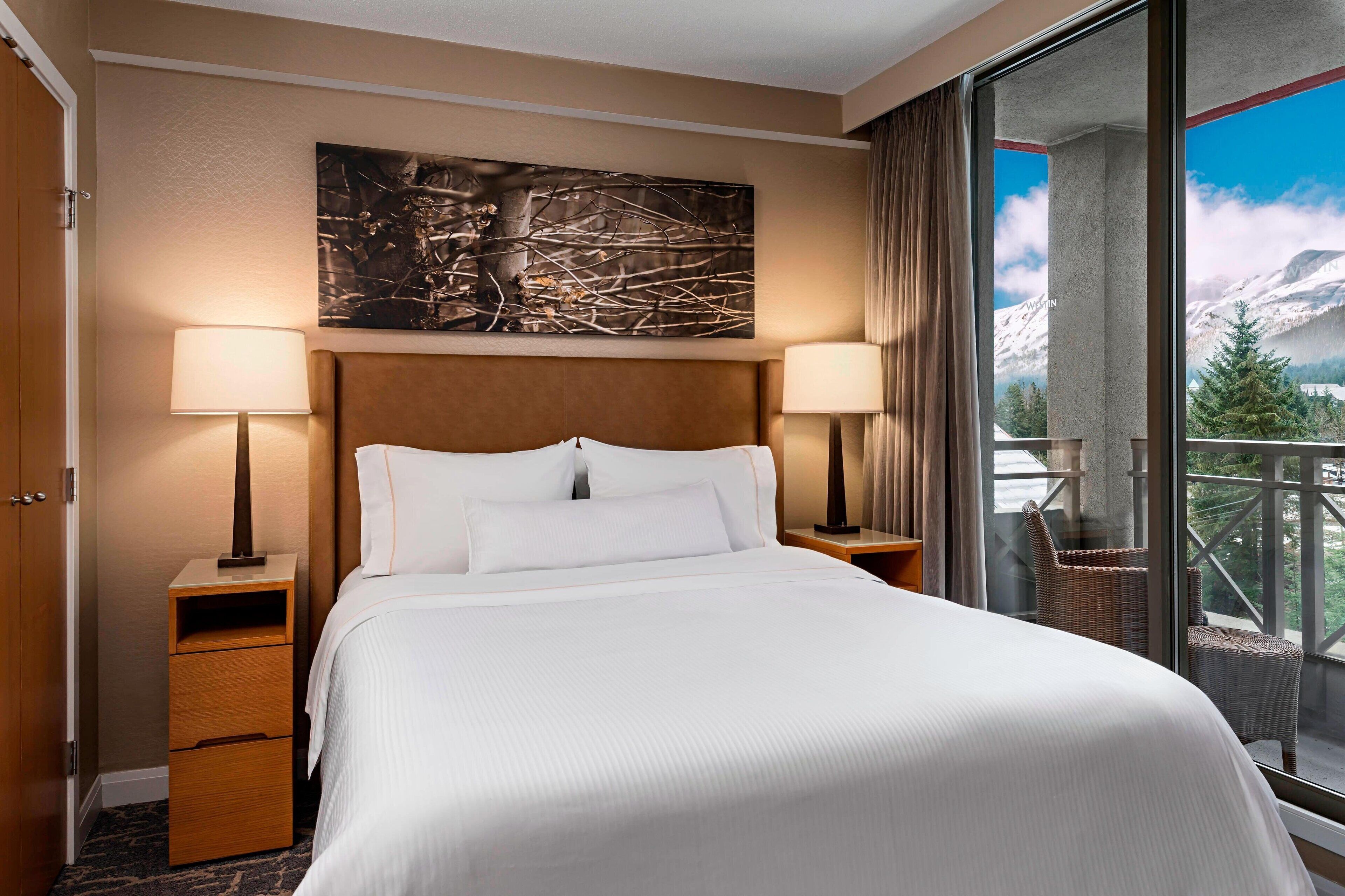 Une chambre d’hôtel avec un grand lit, deux tables de chevet, une œuvre d’art accrochée au mur et un balcon offrant une vue sur la montagne.