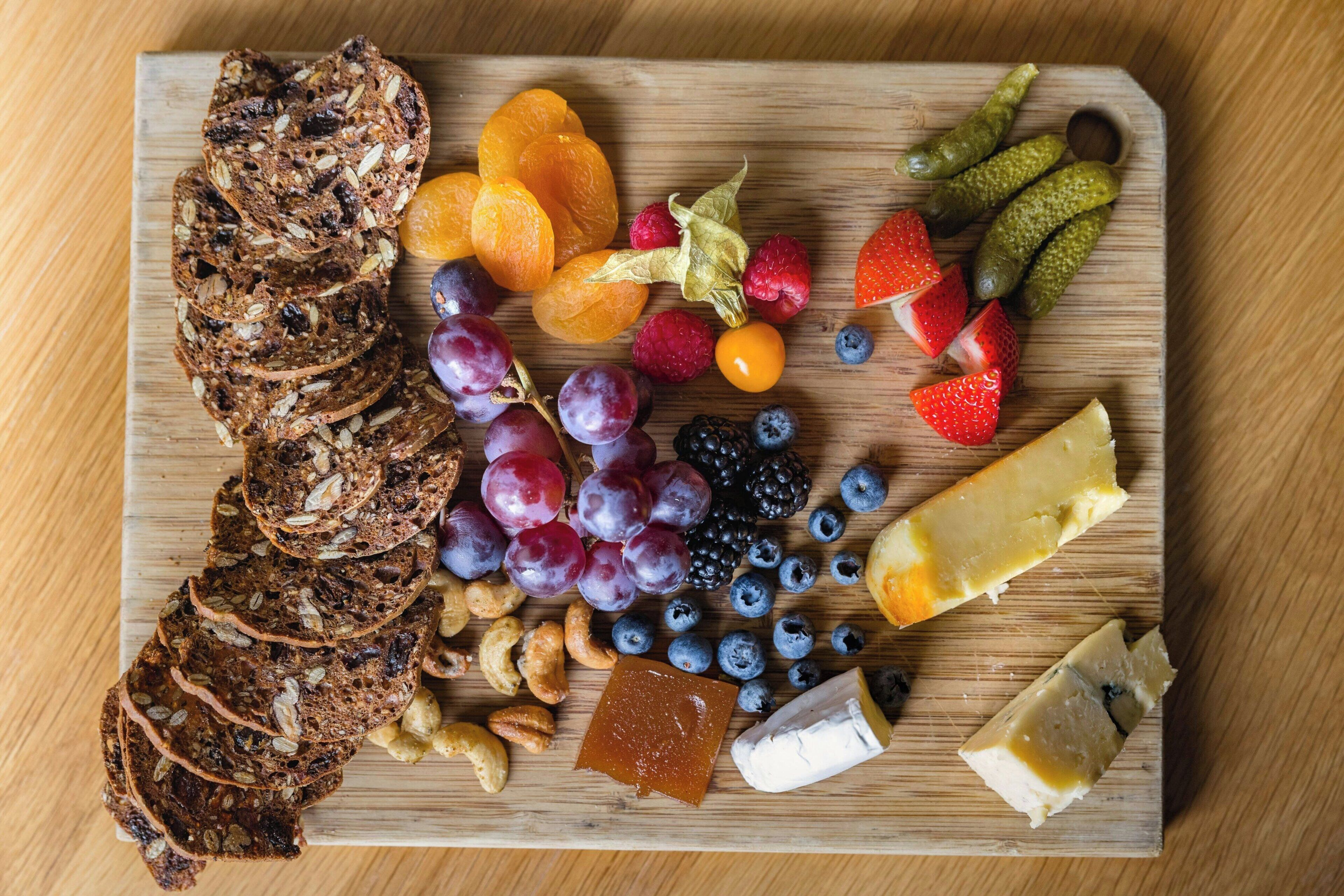 A wooden board with sliced bread, fruits, nuts, and cheese.