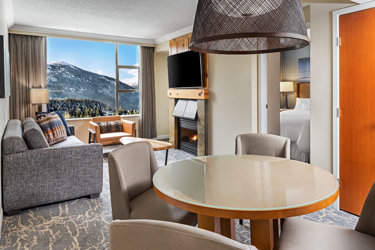 A hotel room with a sofa, chairs, a glass table, a fireplace, a TV, and a mountain view.