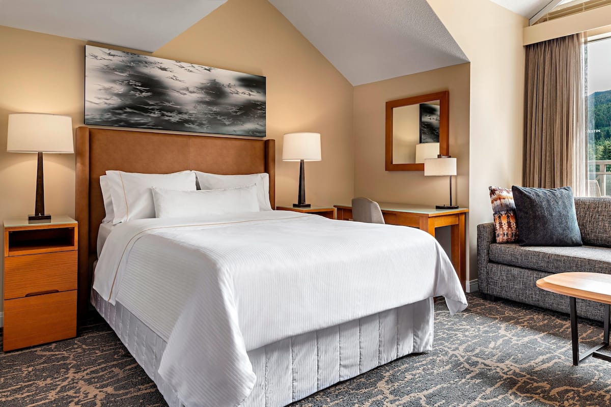 A hotel room with a large bed, two bedside tables, a desk, a chair, a sofa, and a large window with a view.
