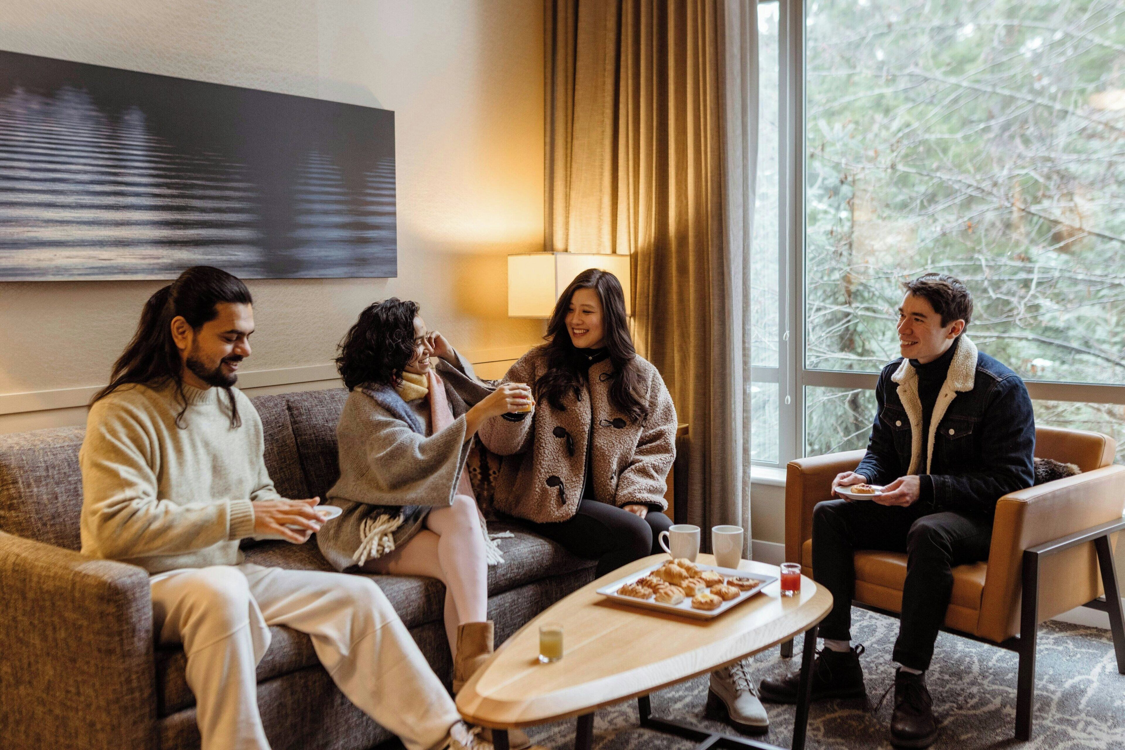 A group of people sitting on a couch and chairs in a room with a large window, a lamp, and a table with food.