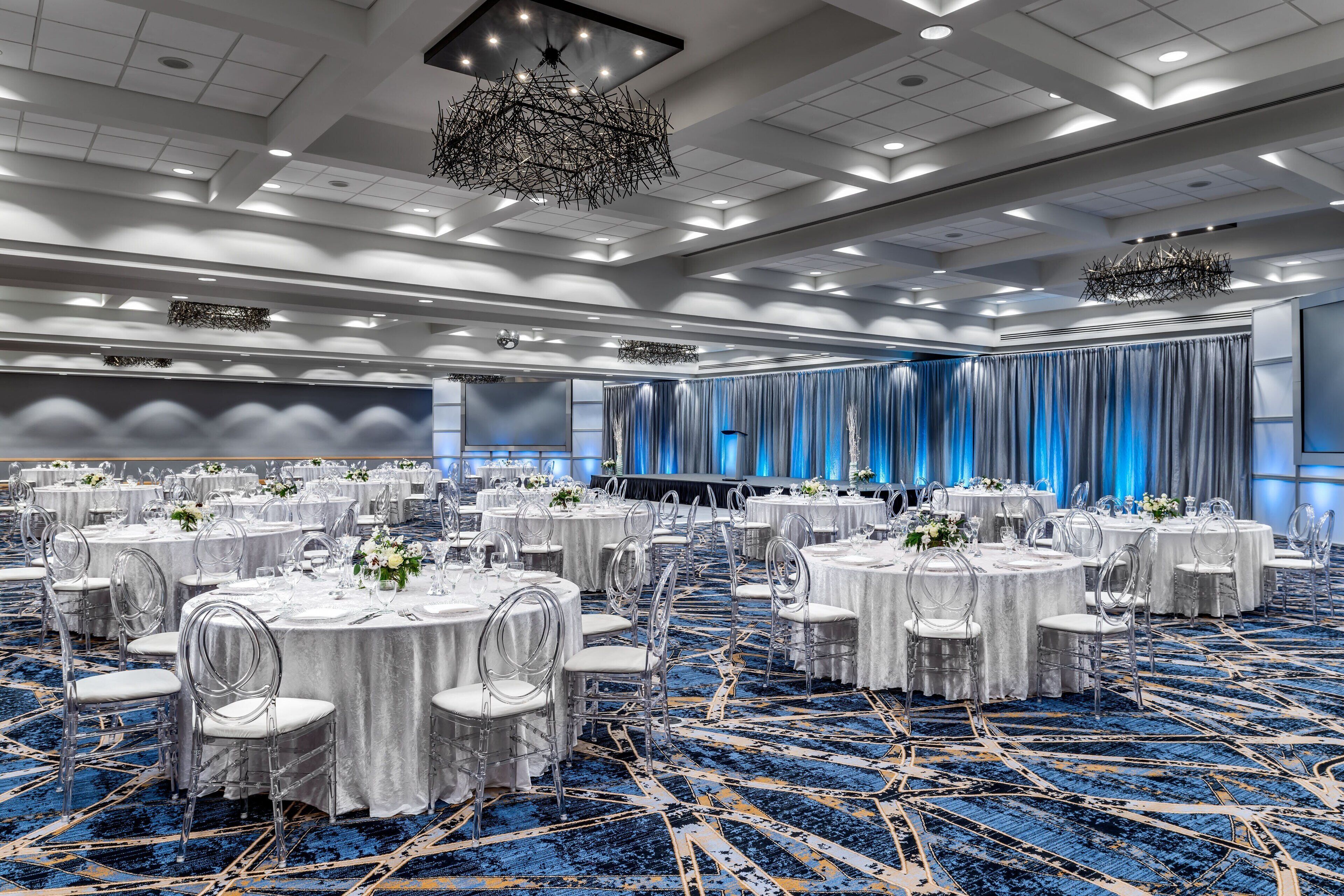 Une salle de banquet avec des tables rondes, des chaises transparentes et une moquette bleue.