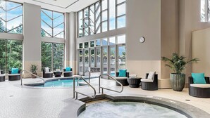 Una amplia piscina interior con varias zonas de estar y un jacuzzi, rodeada de grandes ventanales con vistas a zonas verdes.