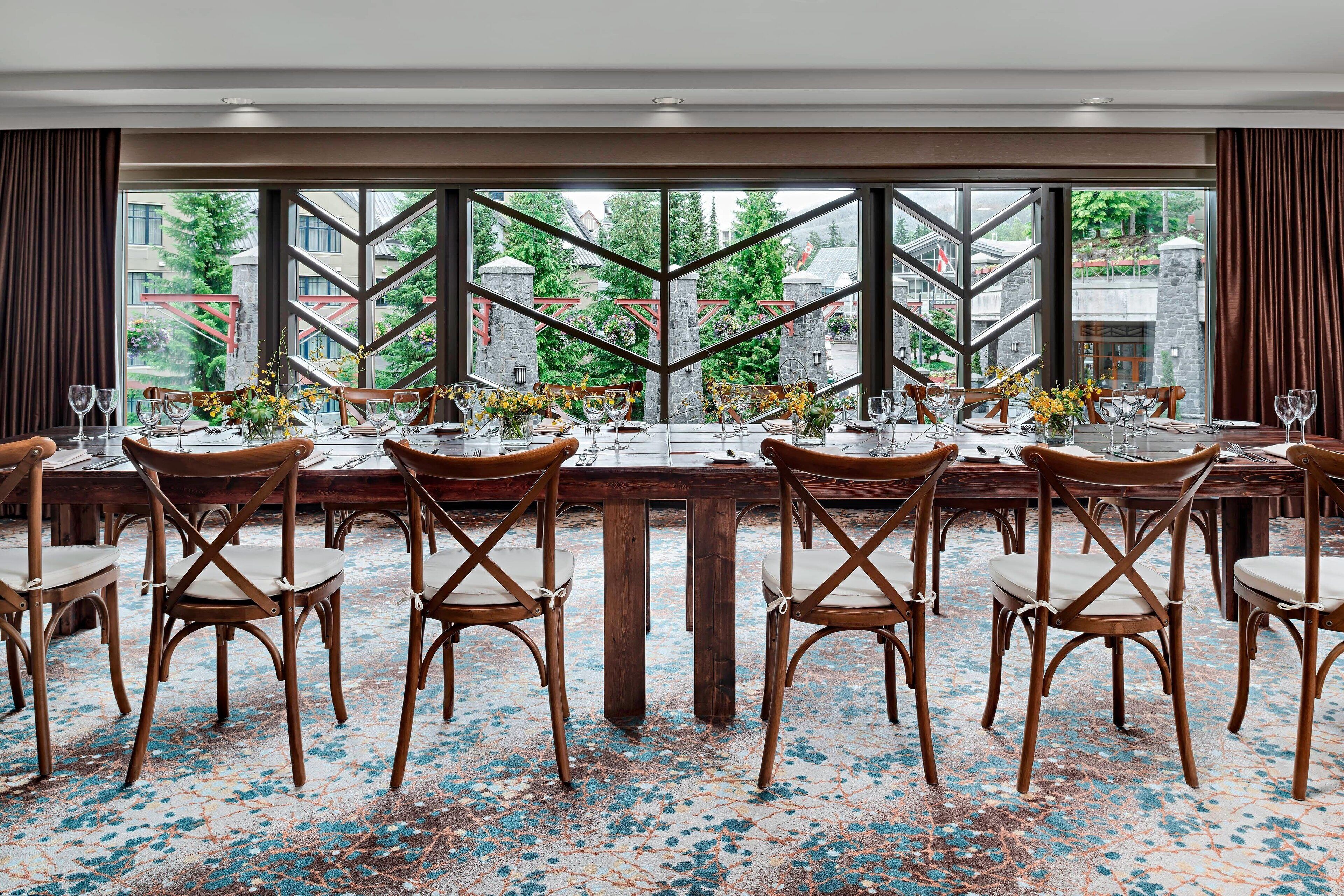 A dining area with a large table set for a meal, surrounded by wooden chairs, and large windows offering a view of buildings and trees.