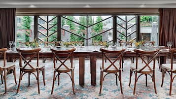 A dining area with a large table set for a meal, surrounded by wooden chairs, and large windows offering a view of buildings and trees.