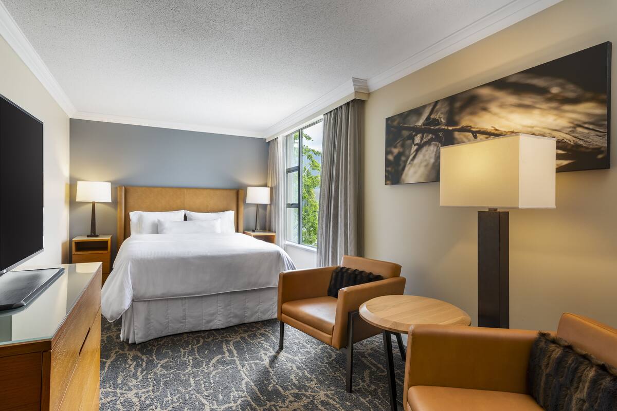 A hotel room with a large bed, a desk with a flat-screen TV, a chair, a small table, and a large window with curtains.