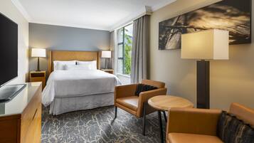 Une chambre d’hôtel avec un grand lit, un bureau doté d’un téléviseur à écran plat, une chaise, une petite table et une grande fenêtre avec des rideaux.