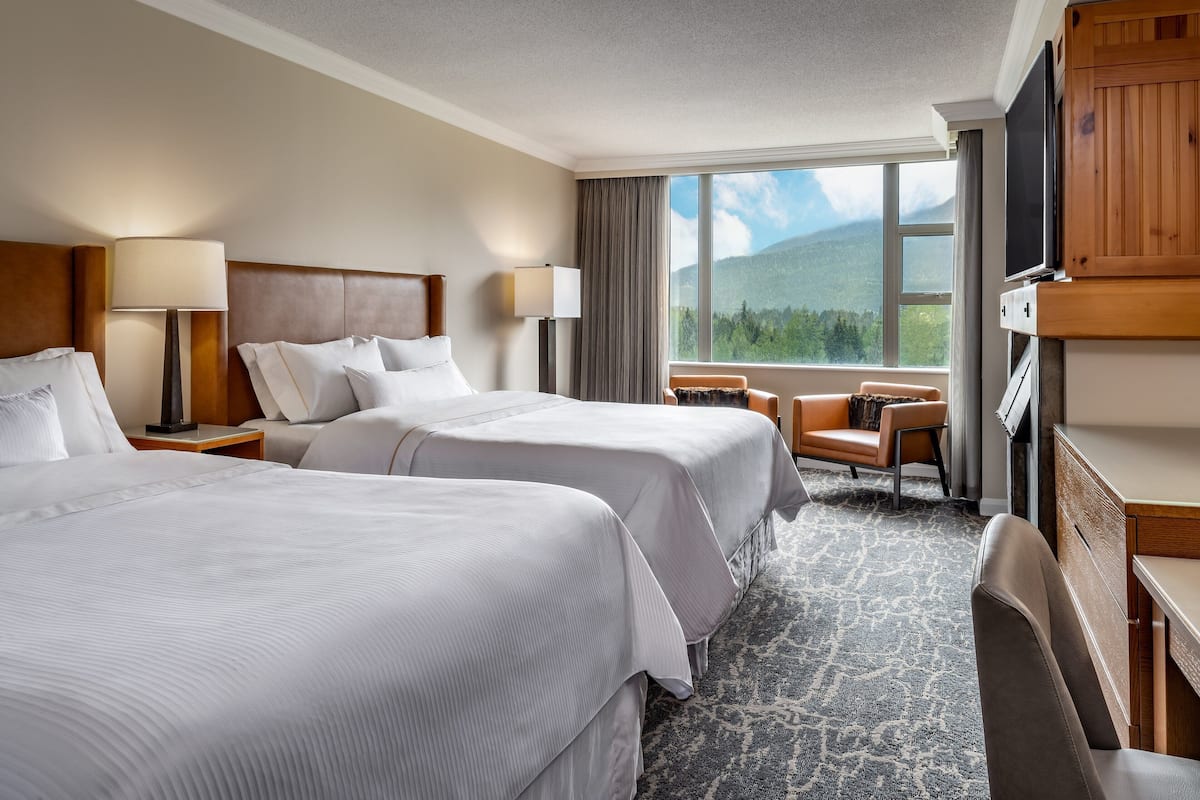 A hotel room with two beds, a desk, a chair, a TV, and a large window with a mountain view.