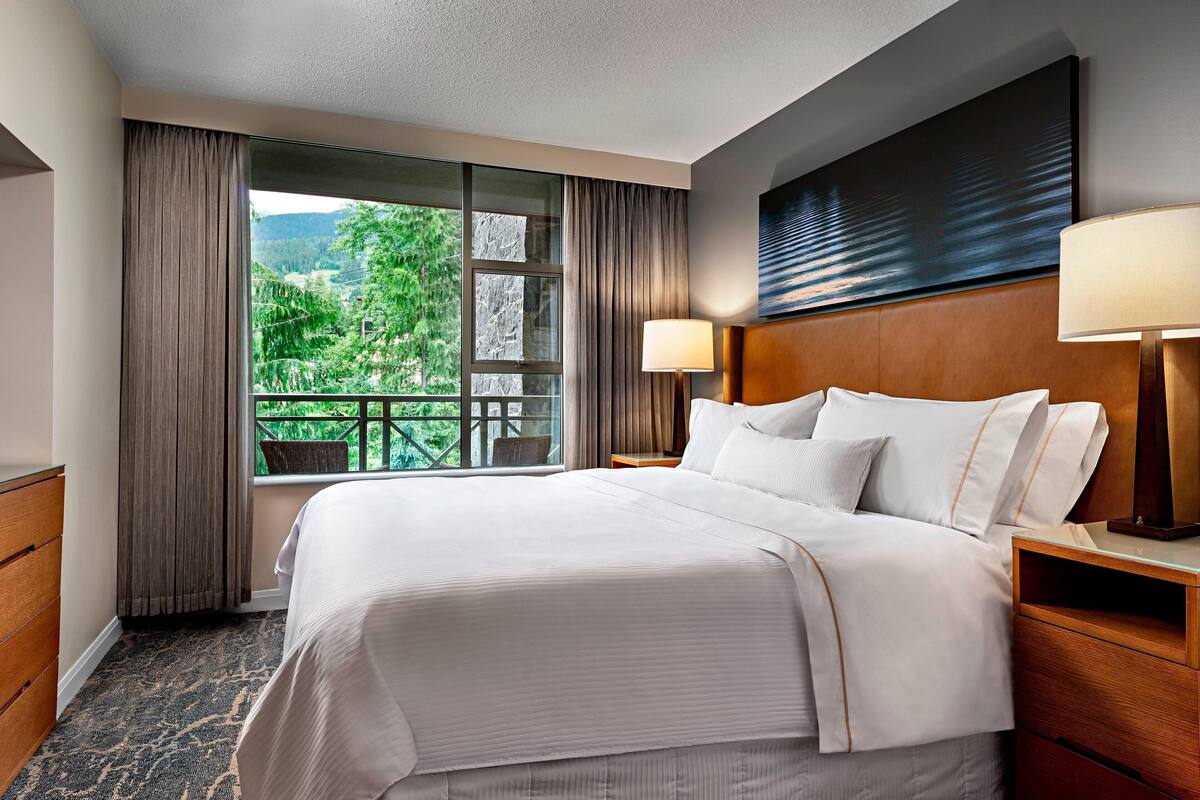 A hotel room with a large bed, two bedside tables, a window with curtains, and a view of greenery outside.