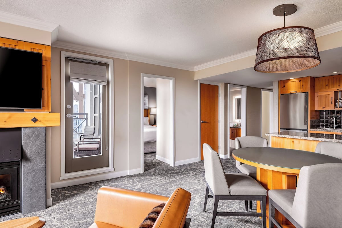 A modern hotel room with a fireplace, a dining area with a round table and chairs, a kitchen with wooden cabinets, and a large TV mounted on the wall.