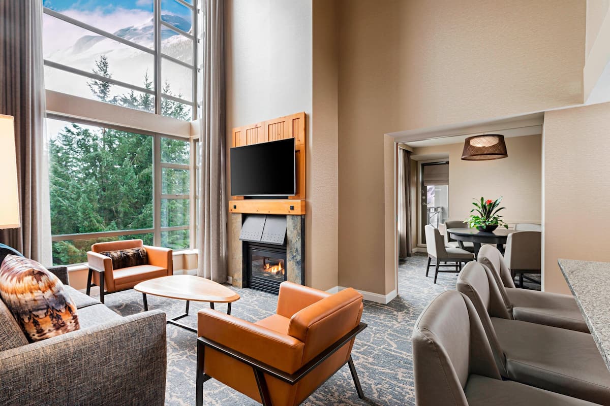 A spacious living area with a fireplace, comfortable seating, a flat-screen TV, and large windows offering a view of trees and mountains.