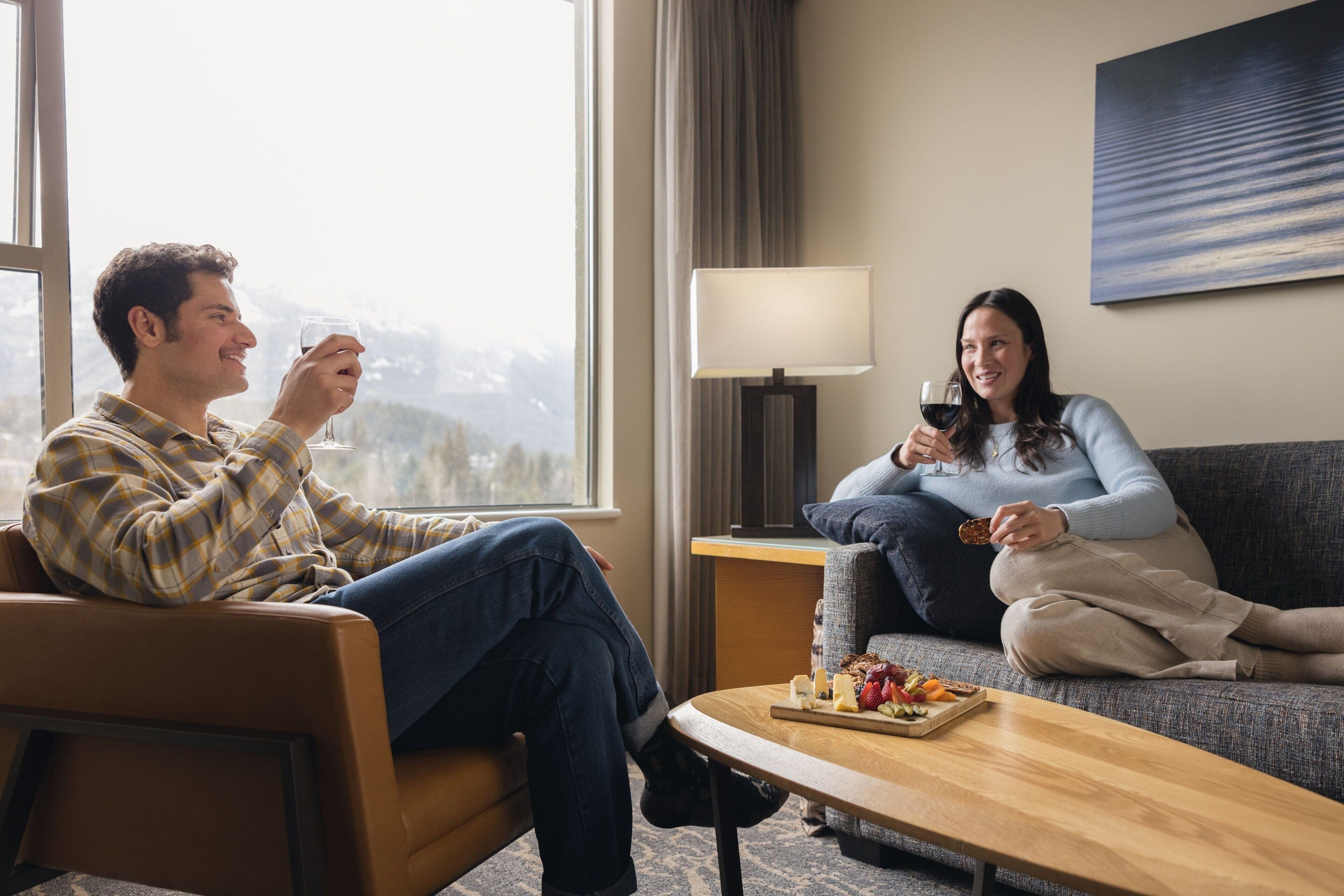 A couple sitting on a couch in a well-lit room with a view of mountains through a large window.