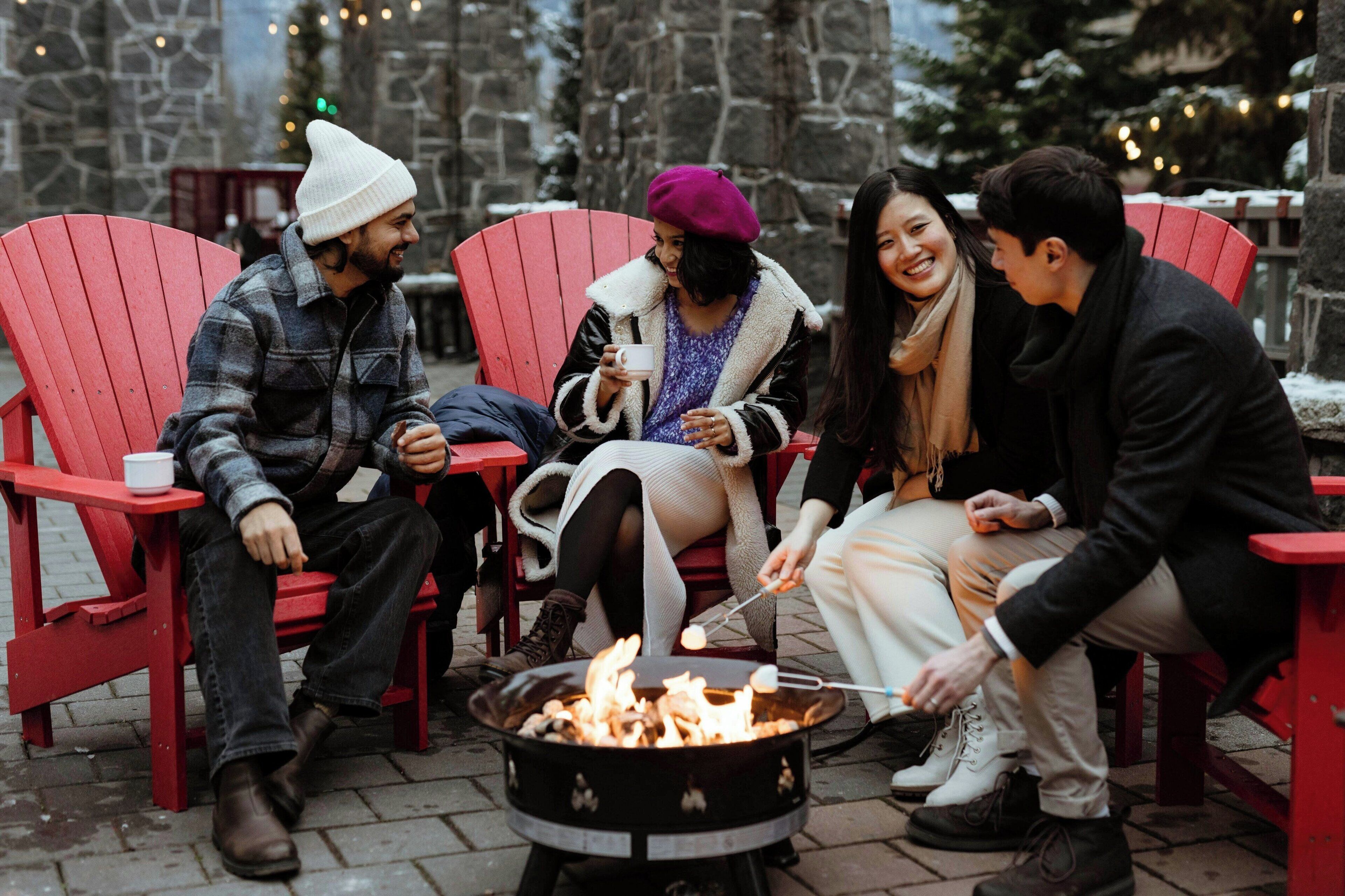 A group of people sitting on red Adirondack chairs around a campfire.
