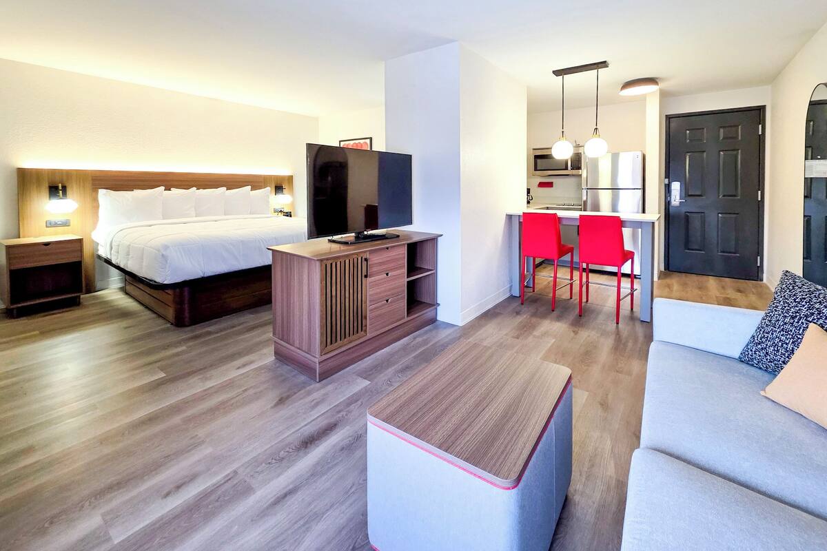 Ein Hotelzimmer mit Bett, Fernseher, einer kleinen Küchenzeile, einem Essbereich mit roten Stühlen und einer Couch.