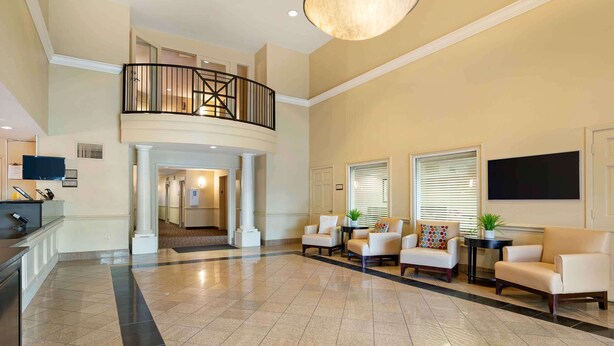 A spacious lobby with a high ceiling, a central seating area with sofas and armchairs, a staircase leading to an upper level, and a flat-screen TV mounted on the wall. - Extended Stay America Suites Houston Med Ctr Braeswood Bl (Houston)