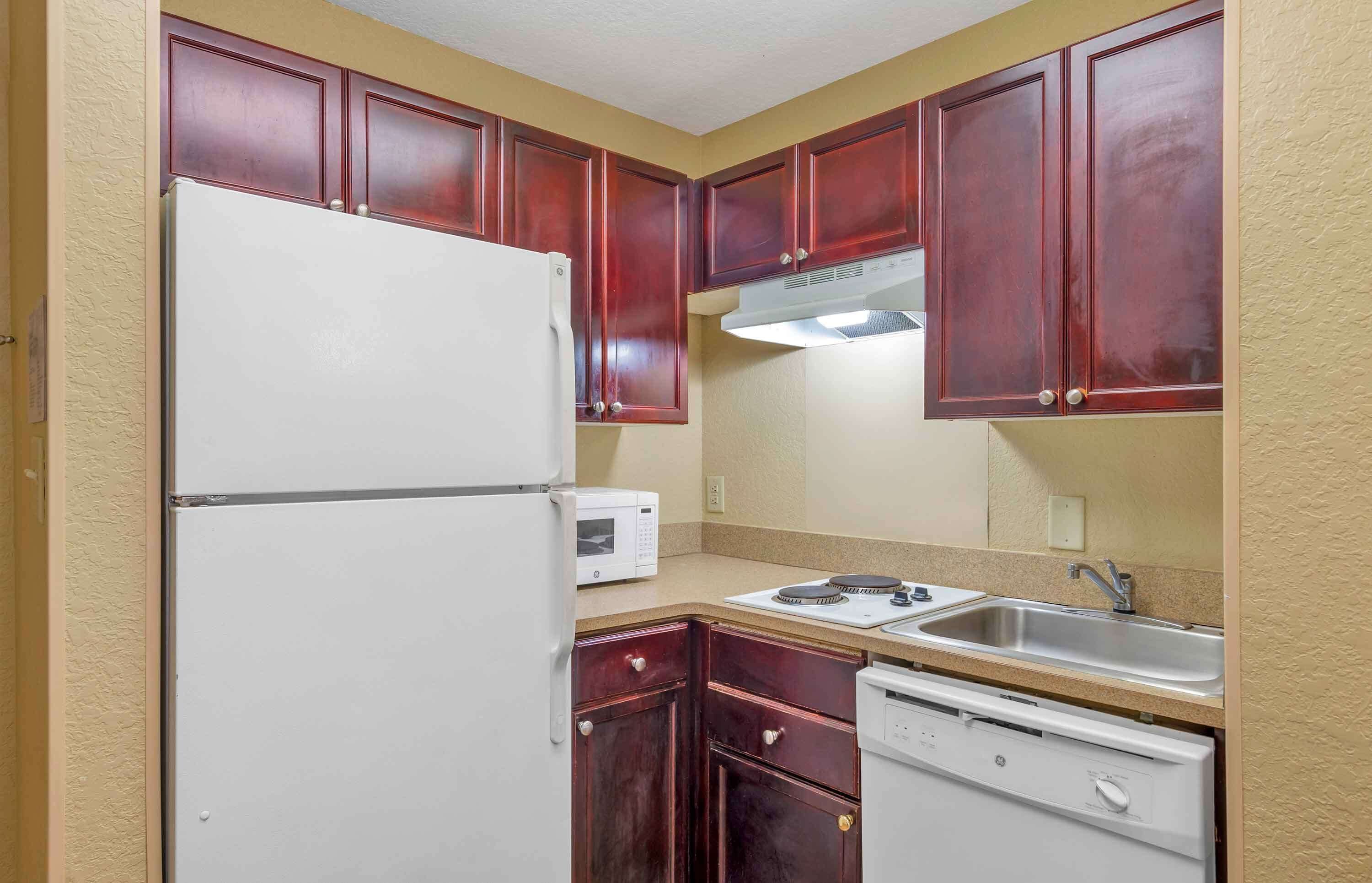 A compact kitchen with wooden cabinets, a white refrigerator, a microwave, a dishwasher, and a stainless steel sink.