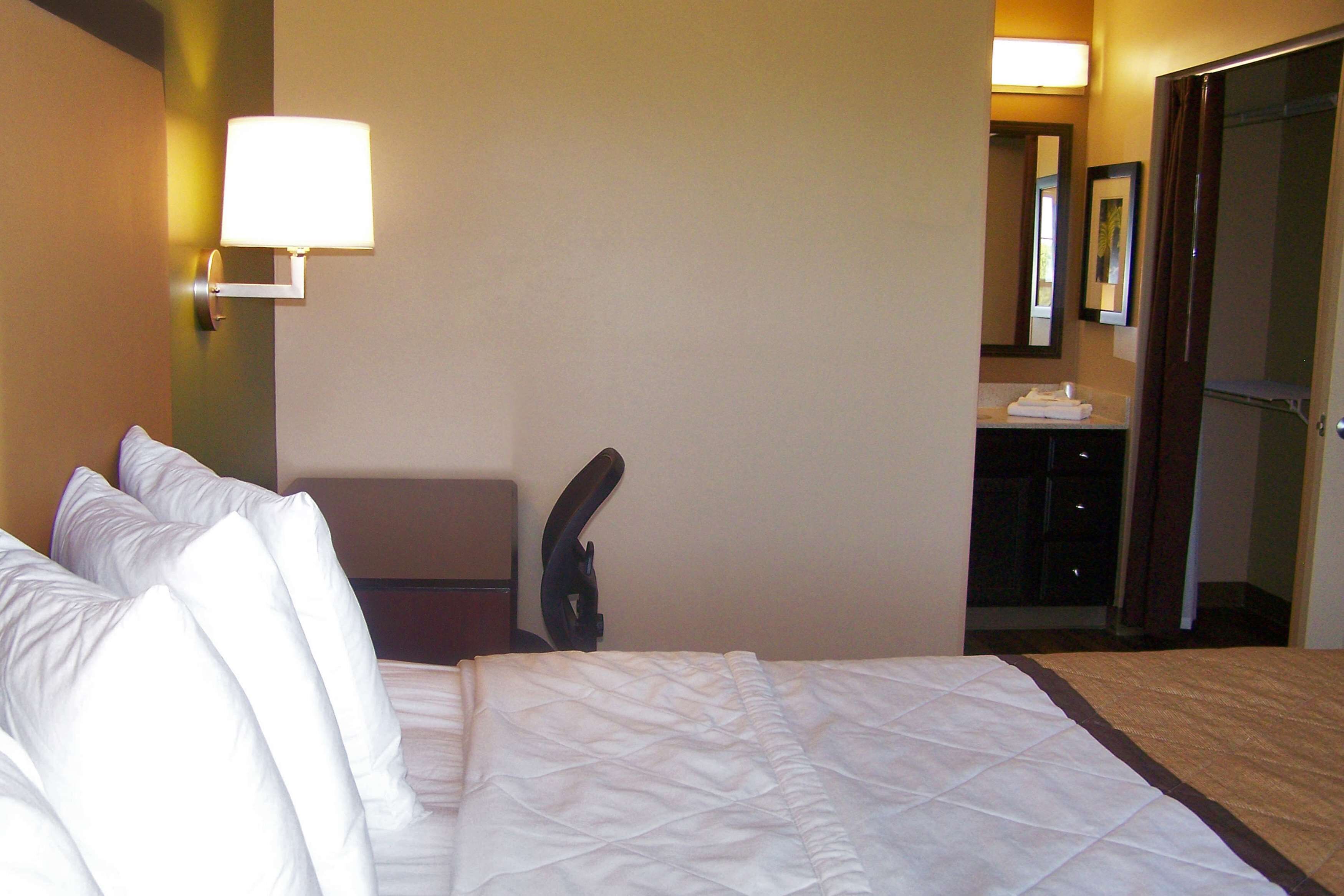 A hotel room with a bed, a desk, a chair, a lamp, and a bathroom visible through an open door.