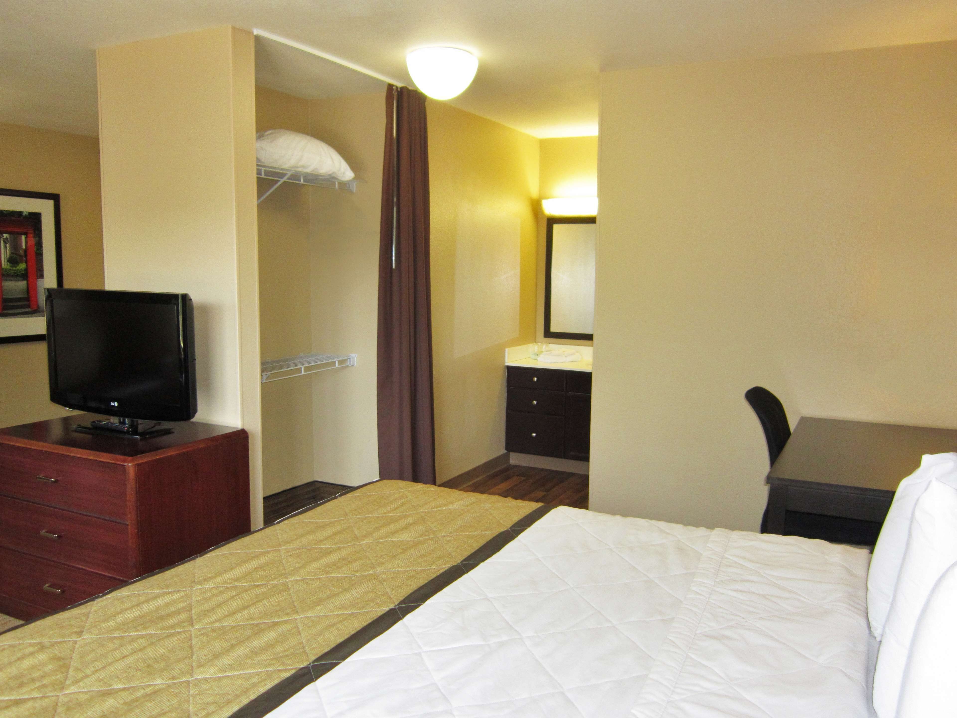 Habitación de hotel con cama, televisión sobre un tocador de madera, baño con lavamanos y espejo, y una silla.