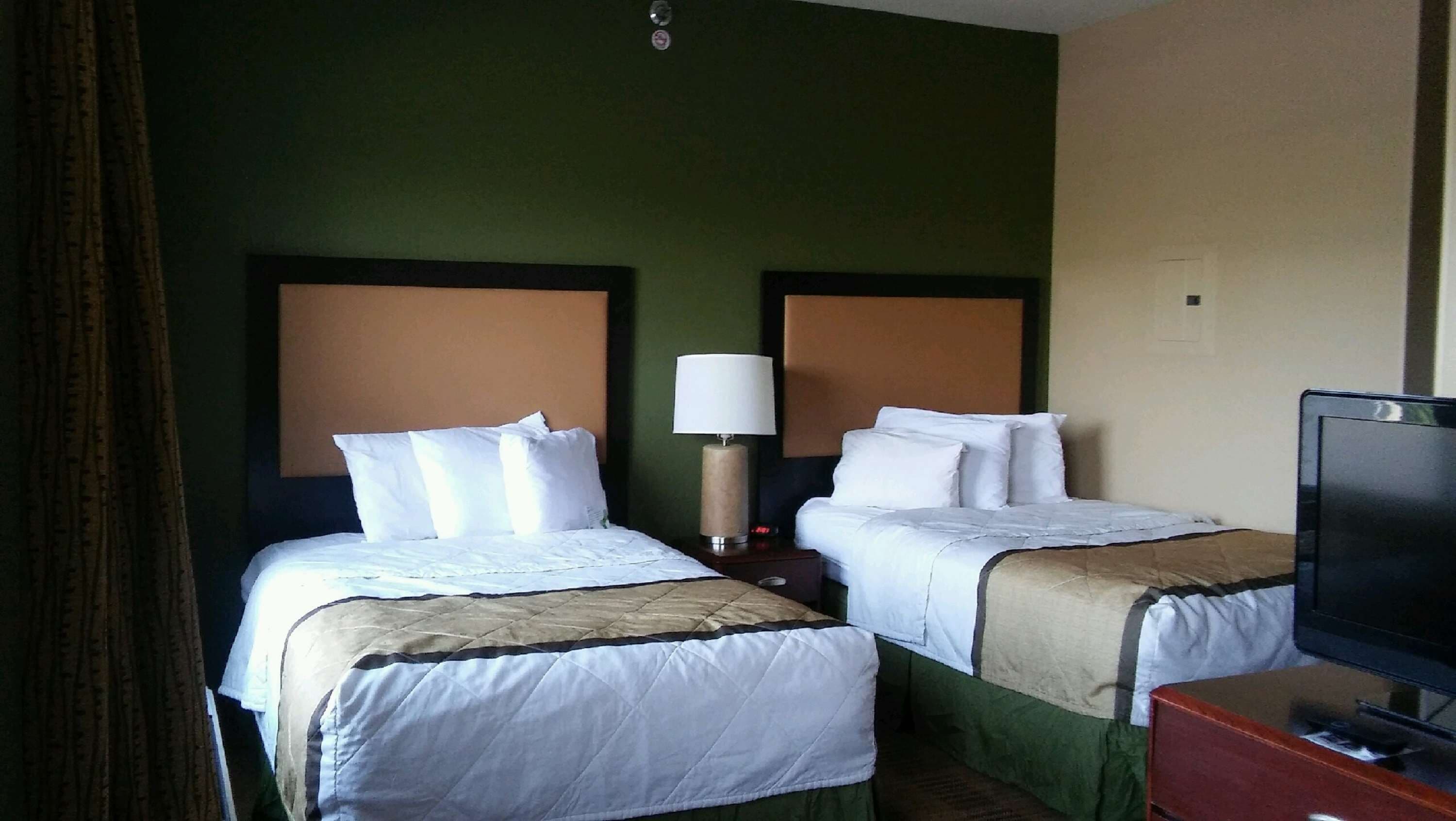 A hotel room with two beds, each with white bedding and a beige headboard. There is a bedside table with a lamp between the beds.