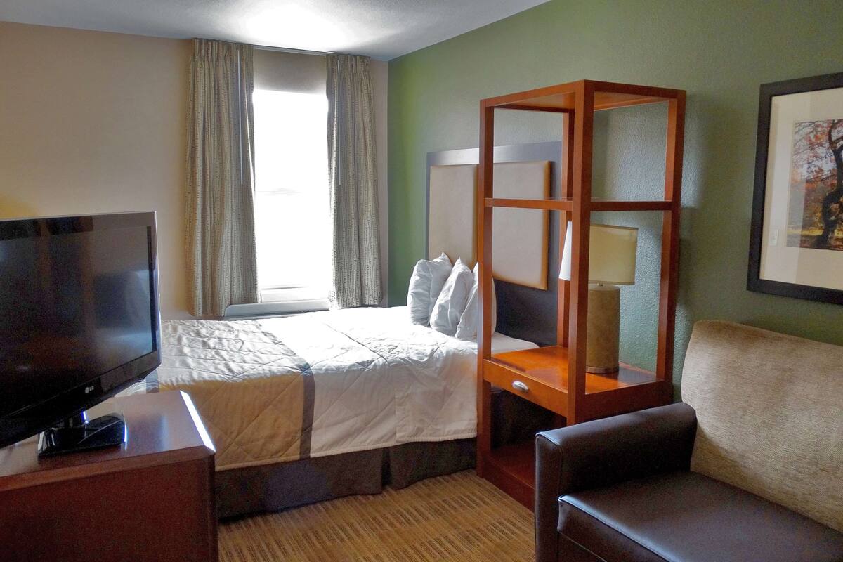 Een hotelkamer met een bed, een televisie, een houten nachtkastje, een stoel en een raam met gordijnen.